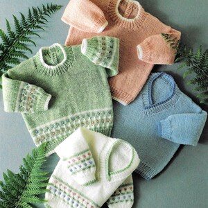 Könnte beinhalten: Vier handgestrickte Pullover in Pastellfarben: Hellrosa, Grün, Blau und Weiß. Jeder Pullover hat ein einzigartiges Design mit gemusterten Bündchen und unteren Rändern. Die Pullover sind auf einer hellblau-grünen Oberfläche angeordnet, mit Farnblättern im Hintergrund.