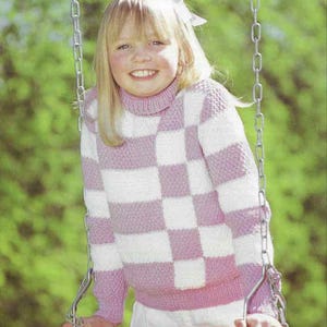 May include: A long-sleeved knitted sweater featuring a checkered pattern in alternating blocks of soft pink and white. The garment is designed with a high turtleneck collar and ribbed cuffs at the wrists and hem, providing a structured fit.