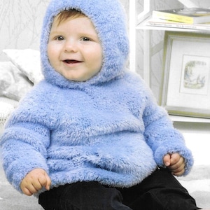 May include: A baby wearing a light blue, fluffy hooded sweater and black pants. The sweater has a soft, textured appearance. The baby is smiling and sitting on a light-colored surface.