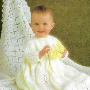 May include: A baby in a white knitted dress and blanket, holding yellow flowers. The blanket has a diamond pattern and yellow accents. The background is a solid green.