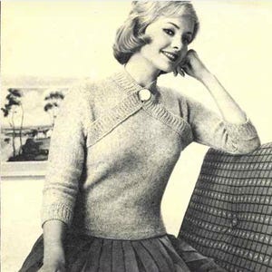 May include: A vintage light gray knitted sweater with a unique cross-over design, featuring a round neckline and three-quarter sleeves. The sweater is fastened with a large, round button. The woman is sitting on a chair with a patterned back.