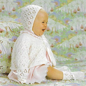 Może przedstawiać: Niemowlę w białym, dzianinowym kapturku, sweterku i skarpetkach. Kapturek i sweterek mają ażurowy wzór. Niemowlę ma również na sobie różową sukienkę z plisowaną spódnicą. Strój uzupełniają białe wstążki.