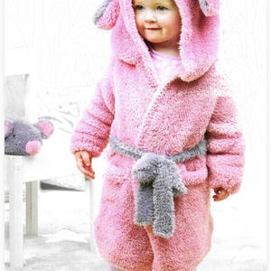 May include: A pink plush hooded robe with gray bunny ears and a matching gray belt. The robe is worn by a child, paired with pink polka dot pajamas. A stuffed animal with bunny ears sits on a white chair.