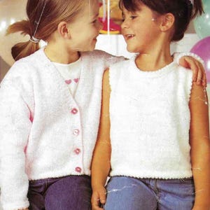 Peut inclure: Deux jeunes filles vêtues de hauts en tricot blanc et de jeans bleus se sourient. L'une porte un cardigan à boutons roses, l'autre un haut sans manches. Des ballons et un ours en peluche sont visibles en arrière-plan.