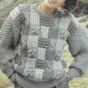 May include: A gray, patchwork-style sweater with a round neckline and long sleeves. The sweater features various textured knit patterns, including cable knit and floral accents. The color palette consists of different shades of gray.