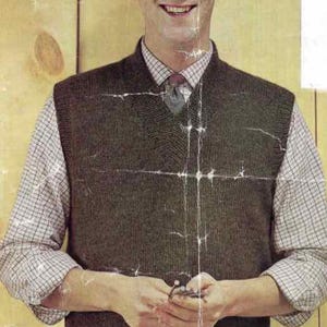 Puede incluir: Un hombre con un chaleco de punto verde oscuro sobre una camisa a cuadros y una corbata. El chaleco tiene un diseño de cuello en V. El fondo es una pared de madera marrón claro.