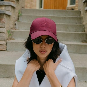 Könnte beinhalten: Eine weinrote Baseballkappe mit kleinem gesticktem Design, goldgerahmte Sonnenbrille und ein weißes ärmelloses Kapuzenshirt. Die Person sitzt auf Betontreppen, trägt schwarze Leggings und ein goldenes Armband.