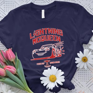 May include: Navy blue t-shirt featuring a red and white graphic of Lightning McQueen from the movie Cars. The design includes the text "LIGHTNING McQUEEN" and "Piston Cup Racing Series". The shirt is surrounded by pink tulips and white daisies.