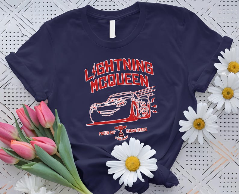 May include: Navy blue t-shirt featuring a red and white graphic of Lightning McQueen from the movie Cars. The design includes the text "LIGHTNING McQUEEN" and "Piston Cup Racing Series". The shirt is surrounded by pink tulips and white daisies.