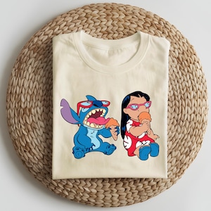 May include: Cream-colored t-shirt featuring a cartoon illustration of Stitch and Lilo from Lilo & Stitch. Stitch is blue, wearing sunglasses, and holding ice cream. Lilo is wearing a red dress and glasses, also holding ice cream.