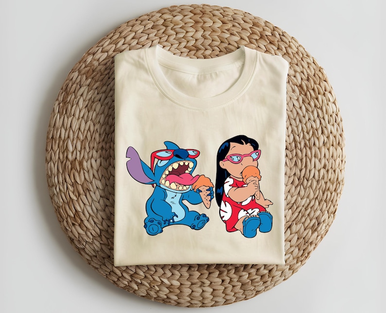 May include: Cream-colored t-shirt featuring a cartoon illustration of Stitch and Lilo from Lilo & Stitch. Stitch is blue, wearing sunglasses, and holding ice cream. Lilo is wearing a red dress and glasses, also holding ice cream.