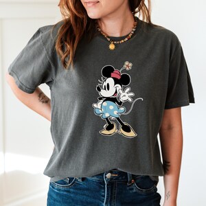May include: A charcoal gray short-sleeve t-shirt featuring a graphic print of Minnie Mouse. The character is depicted wearing a pink hat, a light blue skirt with white polka dots, and tan shoes, with a small flower accessory on her head.