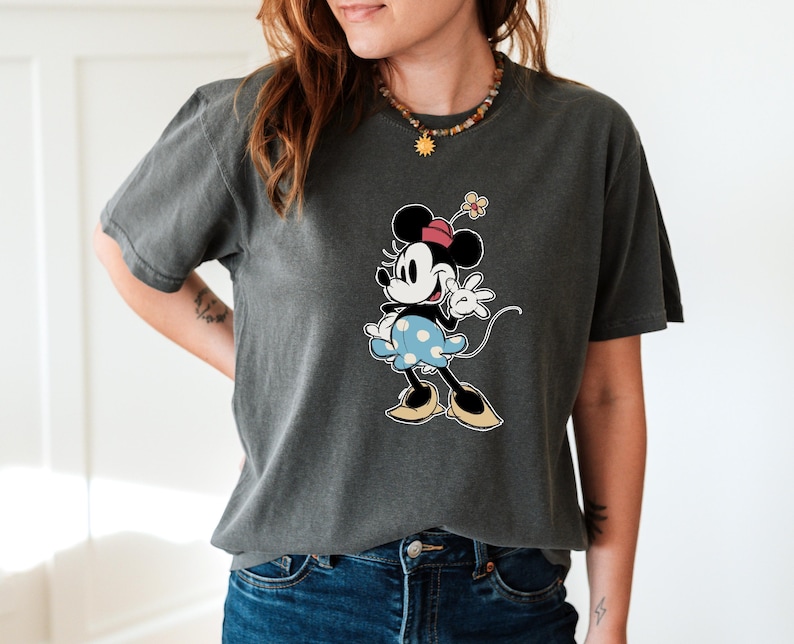 May include: A charcoal gray short-sleeve t-shirt featuring a graphic print of Minnie Mouse. The character is depicted wearing a pink hat, a light blue skirt with white polka dots, and tan shoes, with a small flower accessory on her head.