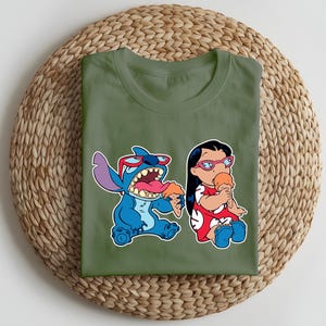 May include: Olive green t-shirt featuring a colorful graphic of Stitch and Lilo from Lilo & Stitch, each holding ice cream cones. Stitch is blue with red sunglasses, and Lilo wears a red dress. The shirt is folded on a woven mat.