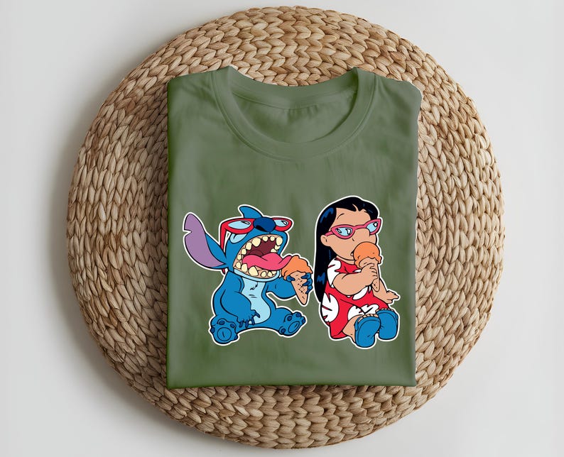 May include: Olive green t-shirt featuring a colorful graphic of Stitch and Lilo from Lilo & Stitch, each holding ice cream cones. Stitch is blue with red sunglasses, and Lilo wears a red dress. The shirt is folded on a woven mat.
