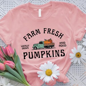 May include: A pale pink t-shirt featuring the text "Farm Fresh Pumpkins" in black. The design includes a green truck laden with pumpkins, with the words "Locally Grown" and "Hand Picked".