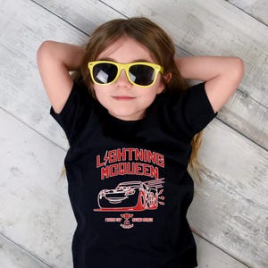 May include: A young person wearing yellow sunglasses and a black t-shirt with a red and white graphic of Lightning McQueen from the Cars movie. The text "LIGHTNING McQUEEN" is above the car graphic.