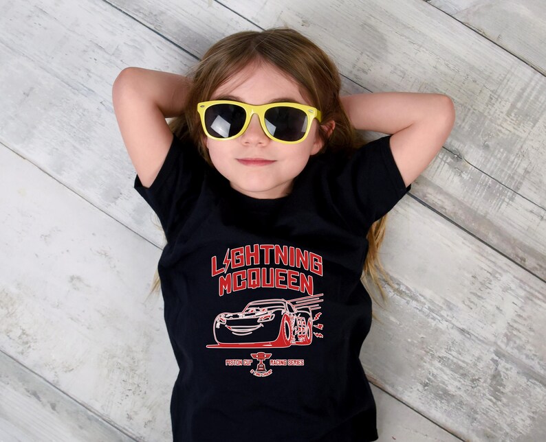 May include: A young person wearing yellow sunglasses and a black t-shirt with a red and white graphic of Lightning McQueen from the Cars movie. The text "LIGHTNING McQUEEN" is above the car graphic.