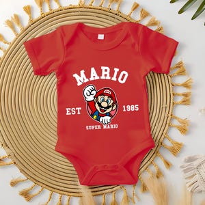 May include: A red baby onesie featuring the text "MARIO" in white arched lettering above a graphic of Mario. The graphic is surrounded by the text "EST 1985 SUPER MARIO". The onesie is short-sleeved.