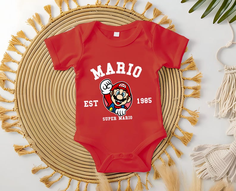 May include: A red baby onesie featuring the text "MARIO" in white arched lettering above a graphic of Mario. The graphic is surrounded by the text "EST 1985 SUPER MARIO". The onesie is short-sleeved.