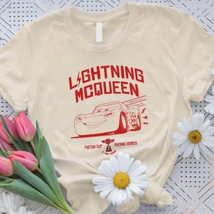 May include: Cream-colored t-shirt featuring a red graphic of Lightning McQueen from the movie Cars. The design includes the text "LIGHTNING MCQUEEN" and "Piston Cup Racing Series".