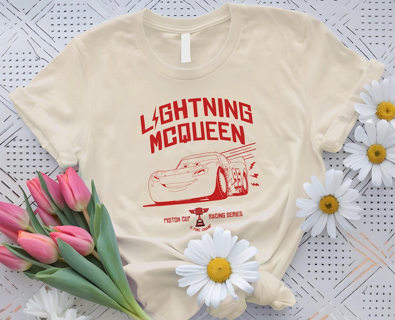 May include: Cream-colored t-shirt featuring a red graphic of Lightning McQueen from the movie Cars. The design includes the text "LIGHTNING MCQUEEN" and "Piston Cup Racing Series".