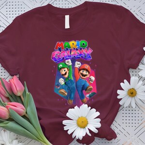 Puede incluir: Camiseta burdeos con un gr&aacute;fico colorido de Mario y Luigi y el texto "MARIO GALAXY". El dise&ntilde;o muestra a los personajes en poses de acci&oacute;n. Las margaritas blancas y los tulipanes rosas rodean la camiseta.