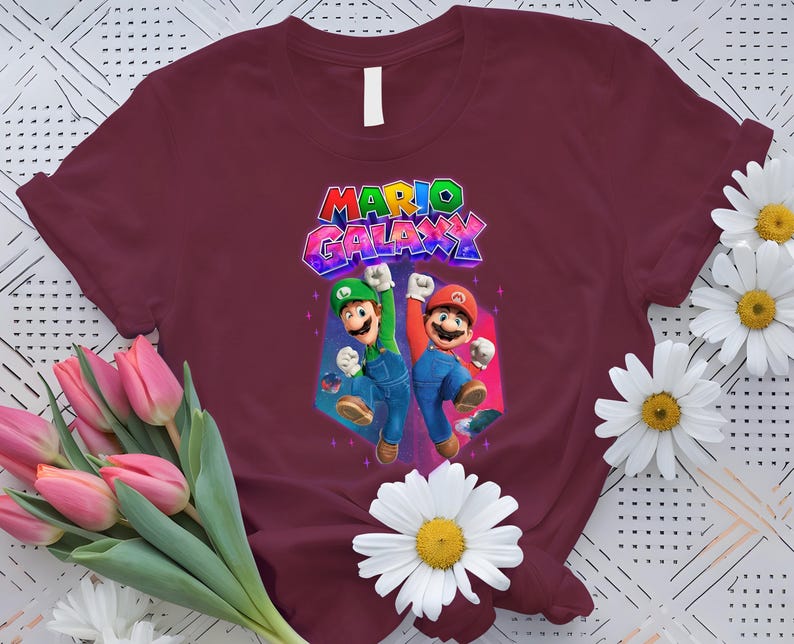 Puede incluir: Camiseta burdeos con un gr&aacute;fico colorido de Mario y Luigi y el texto "MARIO GALAXY". El dise&ntilde;o muestra a los personajes en poses de acci&oacute;n. Las margaritas blancas y los tulipanes rosas rodean la camiseta.