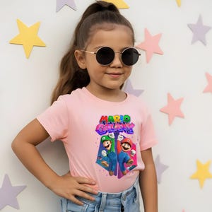 Puede incluir: Una persona joven con una camiseta rosa claro con las palabras "MARIO GALAXY" en una fuente 3D colorida. La camiseta presenta un gr&aacute;fico de Mario y Luigi. La persona lleva gafas de sol y vaqueros azules.