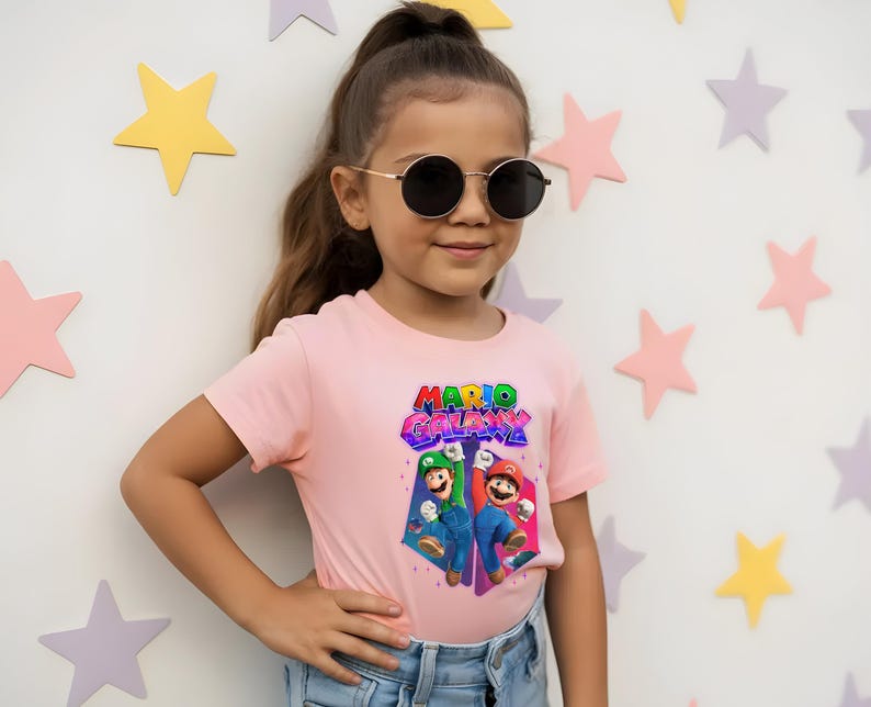 Puede incluir: Una persona joven con una camiseta rosa claro con las palabras "MARIO GALAXY" en una fuente 3D colorida. La camiseta presenta un gr&aacute;fico de Mario y Luigi. La persona lleva gafas de sol y vaqueros azules.