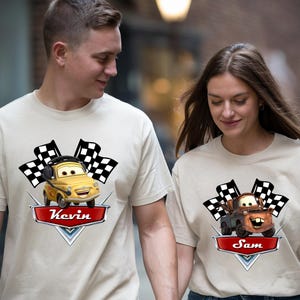 May include: Beige t-shirts featuring cartoon car graphics with checkered flags. One shirt has a yellow car with the name "Kevin" on a red banner. The other shirt has a brown car with the name "Sam".