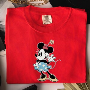 May include: A bright red short-sleeved t-shirt featuring a graphic of Minnie Mouse wearing a pink hat, a light blue polka-dot skirt, and tan shoes. The shirt has a Comfort Colors brand label at the inner neckline with the size XL-TG-EG printed on it.
