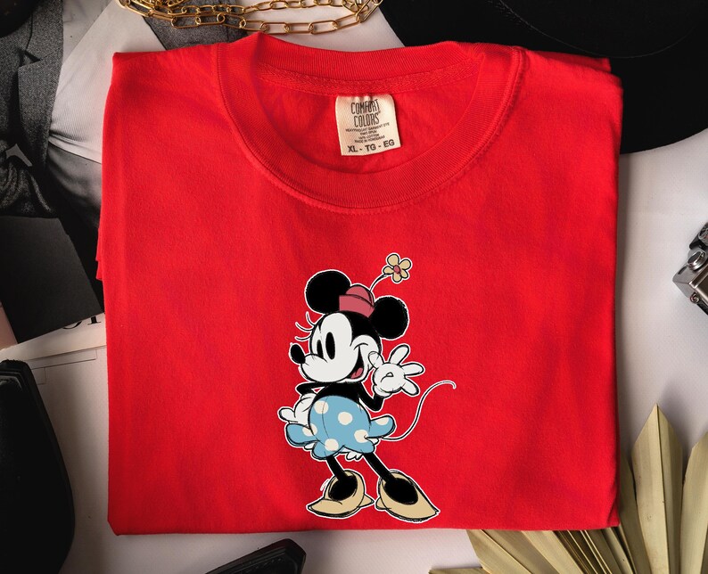 May include: A bright red short-sleeved t-shirt featuring a graphic of Minnie Mouse wearing a pink hat, a light blue polka-dot skirt, and tan shoes. The shirt has a Comfort Colors brand label at the inner neckline with the size XL-TG-EG printed on it.