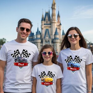 May include: Three people wearing white t-shirts with car graphics and text. The shirts feature a checkered flag design and the words "Dad", "Mom", and "Ellie" on red banners. The background includes a castle.