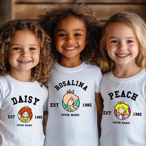 May include: Three young girls wearing white t-shirts featuring Super Mario characters Daisy, Rosalina, and Peach. Each shirt has the character's name in a black arch, the year 1985, and a colorful graphic of the character. The shirts are short-sleeved.