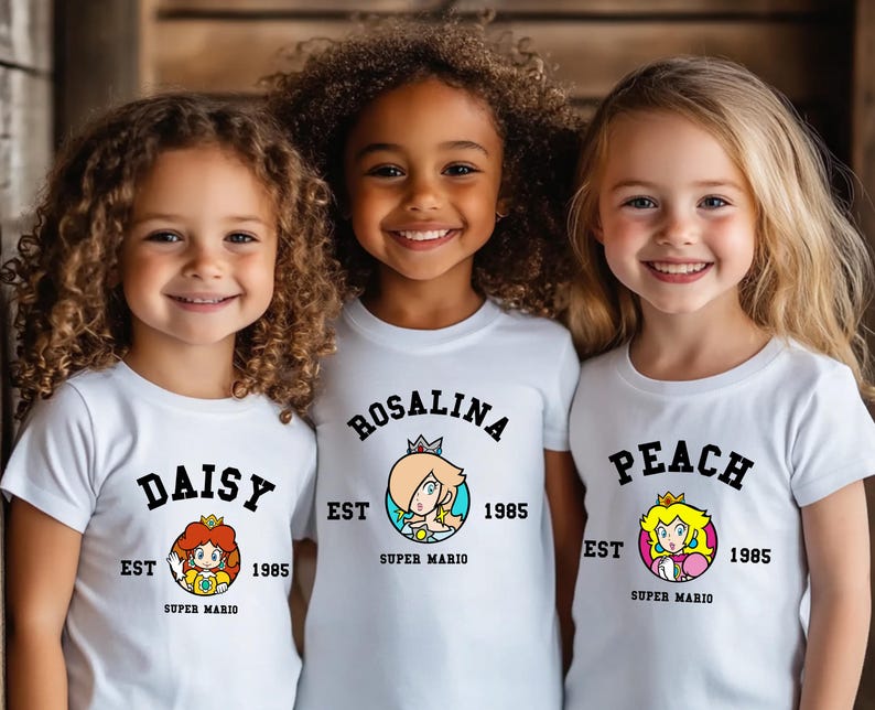 May include: Three young girls wearing white t-shirts featuring Super Mario characters Daisy, Rosalina, and Peach. Each shirt has the character's name in a black arch, the year 1985, and a colorful graphic of the character. The shirts are short-sleeved.