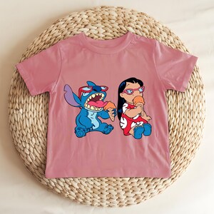 May include: A dusty rose t-shirt featuring a cartoon graphic of Stitch and Lilo eating ice cream cones. Stitch is blue, wearing sunglasses, and Lilo is in a red dress. The shirt is displayed on a woven, circular surface.