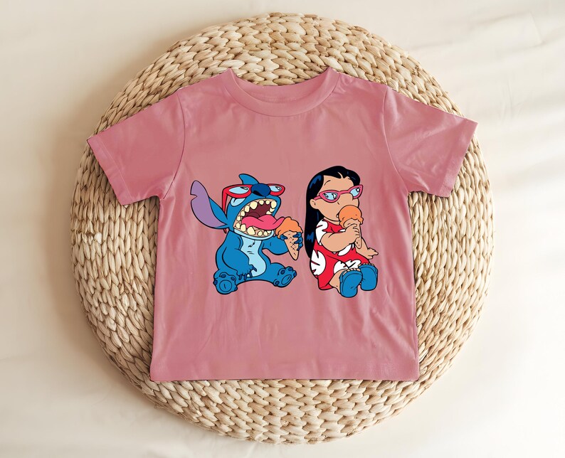 May include: A dusty rose t-shirt featuring a cartoon graphic of Stitch and Lilo eating ice cream cones. Stitch is blue, wearing sunglasses, and Lilo is in a red dress. The shirt is displayed on a woven, circular surface.
