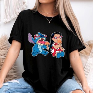 May include: Black t-shirt featuring a cartoon graphic of Stitch and Lilo from the Disney movie Lilo & Stitch. Stitch is blue, wearing sunglasses, and eating ice cream. Lilo is wearing a red dress and eating ice cream.