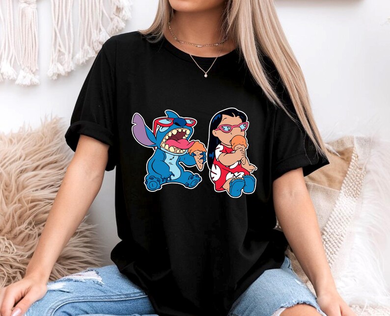 May include: Black t-shirt featuring a cartoon graphic of Stitch and Lilo from the Disney movie Lilo & Stitch. Stitch is blue, wearing sunglasses, and eating ice cream. Lilo is wearing a red dress and eating ice cream.