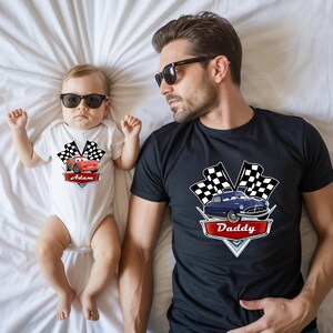 May include: A white baby onesie with a red race car graphic and the name "Adam". A black t-shirt with a blue car graphic and the word "Daddy". Both shirts have checkered flags.