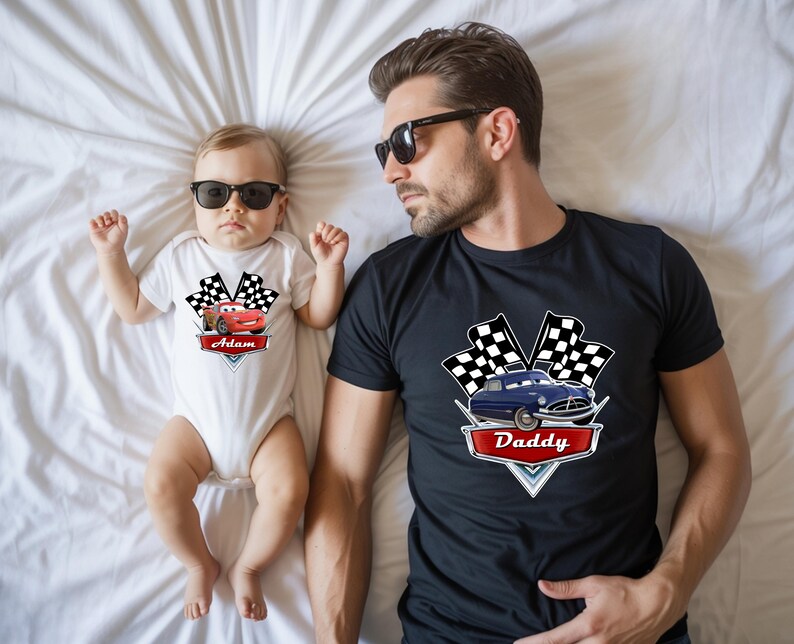 May include: A white baby onesie with a red race car graphic and the name "Adam". A black t-shirt with a blue car graphic and the word "Daddy". Both shirts have checkered flags.