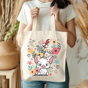 Może przedstawiać: Kremowa torba typu tote z kolorową ilustracją świni i koguta z kresk&oacute;wek otoczonych kwiatami. Torba ma dwa beżowe uchwyty i jest trzymana przez osobę.