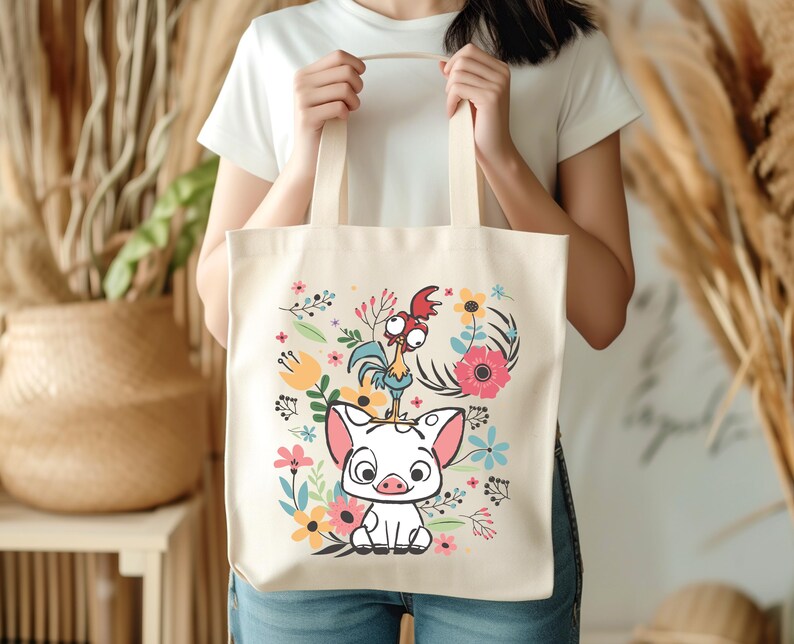 Może przedstawiać: Kremowa torba typu tote z kolorową ilustracją świni i koguta z kresk&oacute;wek otoczonych kwiatami. Torba ma dwa beżowe uchwyty i jest trzymana przez osobę.