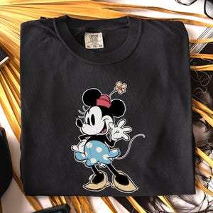 May include: A black crew neck t-shirt featuring a graphic of Minnie Mouse wearing a pink hat with a yellow flower and a light blue skirt with white polka dots. The shirt has a Comfort Colors brand label at the inner neckline showing size XL-TG-EG.