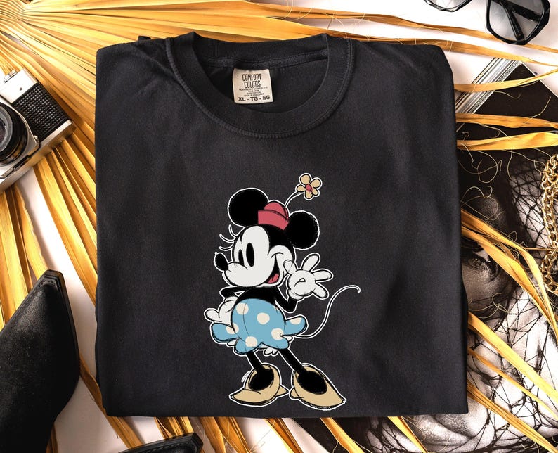 May include: A black crew neck t-shirt featuring a graphic of Minnie Mouse wearing a pink hat with a yellow flower and a light blue skirt with white polka dots. The shirt has a Comfort Colors brand label at the inner neckline showing size XL-TG-EG.