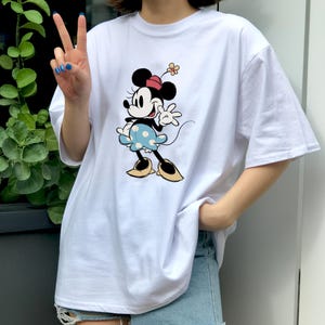 May include: A white short-sleeved t-shirt featuring a graphic print of Minnie Mouse. The character is depicted wearing a red hat with a small flower, a light blue skirt with white polka dots, and yellow shoes, while waving one hand.