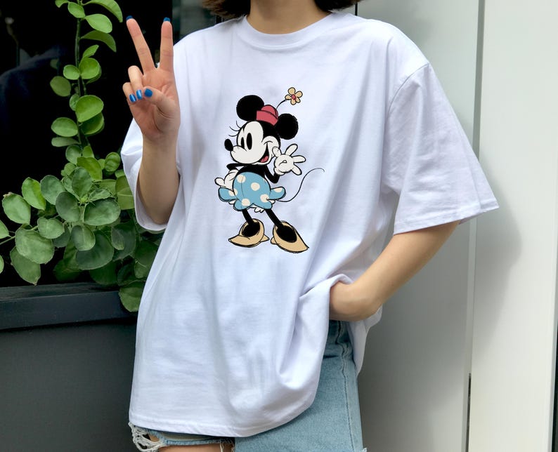 May include: A white short-sleeved t-shirt featuring a graphic print of Minnie Mouse. The character is depicted wearing a red hat with a small flower, a light blue skirt with white polka dots, and yellow shoes, while waving one hand.