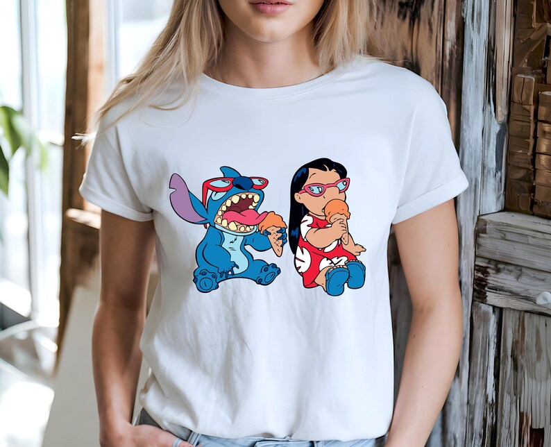 May include: White t-shirt featuring a cartoon illustration of Stitch and Lilo eating ice cream cones. Stitch is blue with red sunglasses, and Lilo wears a red dress and pink glasses. The design is centered on the shirt.