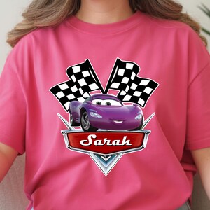 May include: A magenta t-shirt featuring a purple cartoon car with two checkered flags. The car has a smiling face and is set against a red banner with the name "Sarah".